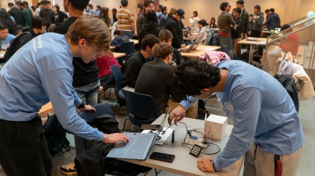 Two builders working on a laptop and breadboard hardware project amid a busy hall.
