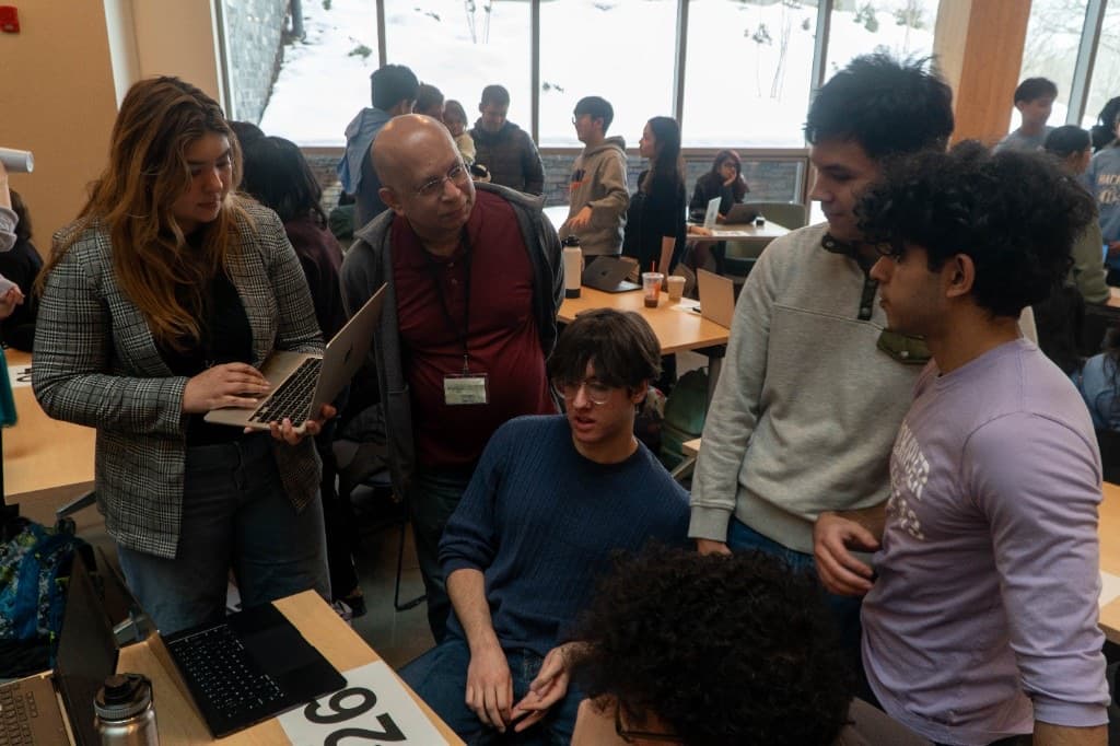 Students and a mentor gathered around laptops at a team table near snowy windows.