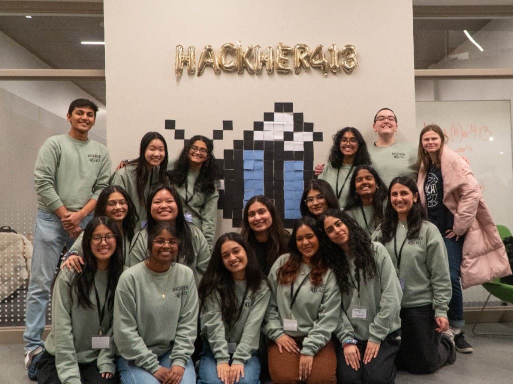 Hack(H)er413 organizers in matching green sweatshirts posing under gold balloon letters and a sticky-note bee.