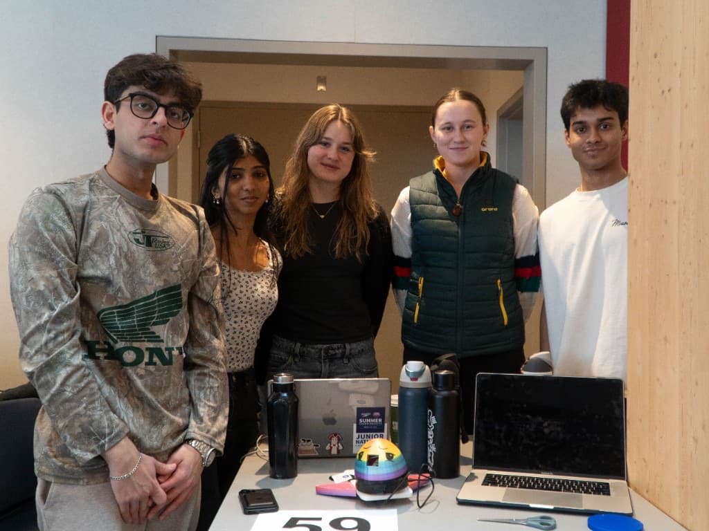 A five-person team posing behind their project table with laptops and a small prototype.